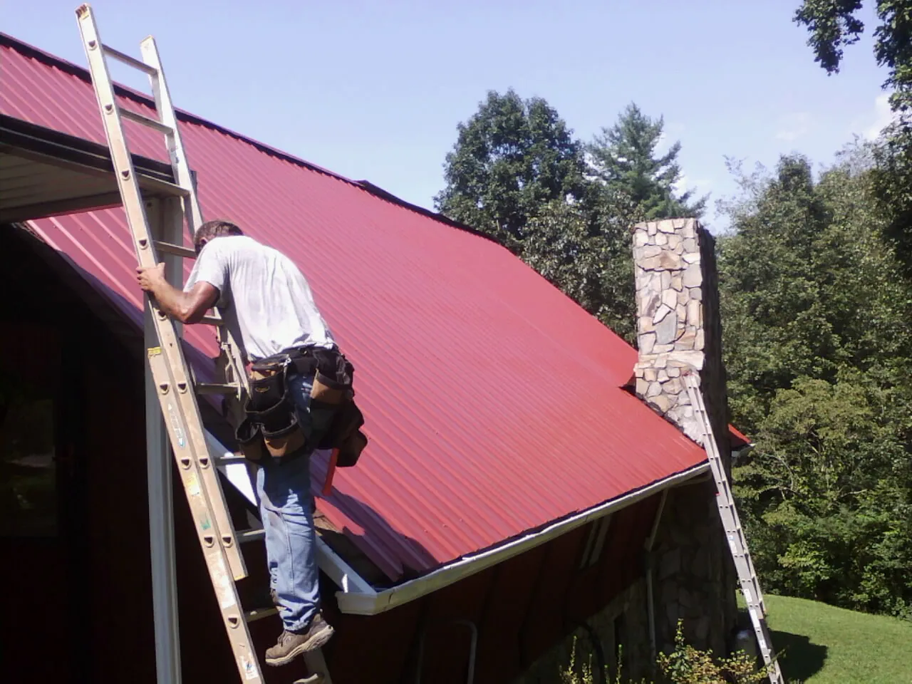 Roofer on a ladder beside a red metal roof and stone chimney — Soffit & Fascia Repair by Roofing Brothers in Dresden